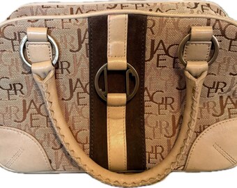 Puede incluir: Bolso de mano color beige con un patrón repetido de las letras "JAC". El bolso presenta una franja central de ante marrón, con un asa y detalles en cuero beige. Anillos metálicos plateados conectan el asa al bolso.
