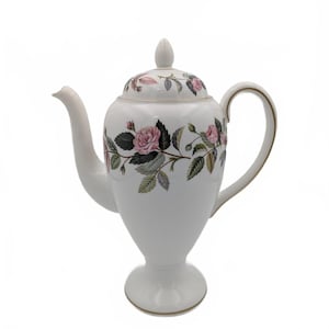 Könnte beinhalten: Weiße Porzellan-Teekanne mit floralem Rosendesign. Die Teekanne hat einen gebogenen Ausguss, einen Henkel und einen Deckel mit Knauf. Rosa Rosen und grüne Blätter verzieren den Körper und den Deckel, mit goldenen Zierakzenten.