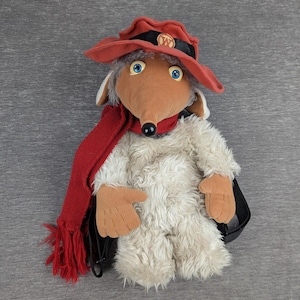 May include: A plush toy character featuring a tan face, blue eyes, and a black nose. It wears a red hat with a black band and a gold W emblem, along with a long red knit scarf. The body is covered in shaggy off-white fur with tan plush hands.