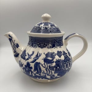 Puede incluir: Una tetera de cerámica blanca con un patrón azul de estilo willow. El diseño incluye ilustraciones detalladas de pagodas, sauces y pájaros en vuelo. La tetera tiene un cuerpo redondeado, un asa curva, un pico vertedor y una tapa a juego con pomo.