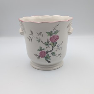 May include: A white ceramic planter with a scalloped rim featuring a thin pink painted edge. The exterior is decorated with a botanical illustration of pink chrysanthemum flowers, green leaves, and dark brown branches. It has two side handles.
