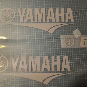 Könnte beinhalten: Zwei Yamaha-Logo-Aufkleber, einer mit dem Markennamen und einem stilisierten Emblem und ein weiterer mit der Zahl 6. Die Aufkleber sind hellfarbig und scheinen aus Vinyl zu sein, vor einem Rasterhintergrund.