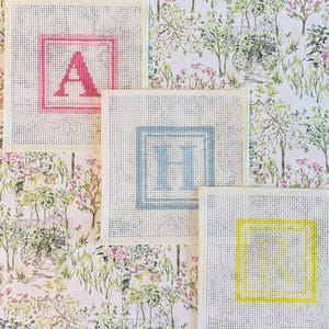 May include: Three square needlepoint canvases featuring centered letters A, H, and R. The letter A is pink, H is light blue, and R is yellow, each enclosed in a matching square border. The canvases are arranged over a floral garden print.