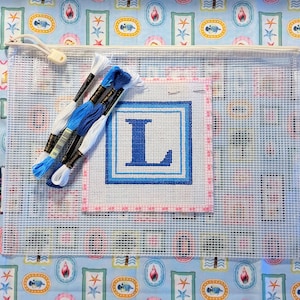 May include: A clear zippered pouch containing a needlepoint kit with the letter "L". The kit includes a canvas with a blue and white design, embroidery floss in shades of blue and white, and a needlepoint canvas. The background is a light blue fabric with a seashell pattern.