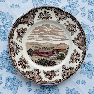 Può includere: Un piatto decorativo vintage con un design paesaggistico marrone e bianco. Il piatto presenta un'illustrazione dettagliata di un edificio e del paesaggio circostante. Il bordo è decorato con motivi floreali e a volute. Il piatto è appoggiato su una tovaglia blu a motivi floreali.