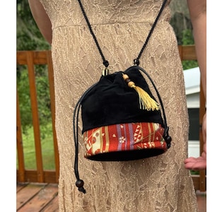 May include: A drawstring bucket bag featuring a black velvet upper section and a lower band with a red, gold, and purple patterned fabric. The bag includes a black cord strap, a yellow tassel accent, and dark wooden beads at the cord ends.