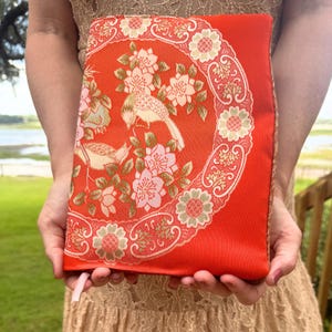 May include: A rectangular fabric book cover in a vibrant orange hue, featuring a woven pattern of light pink and cream-colored flowers, leaves, and two birds. The design is framed by a circular border of stylized floral motifs.