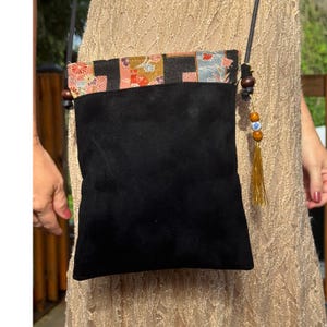 Puede incluir: Un bolso bandolera de ante negro con un panel superior rectangular que presenta un patrón floral y geométrico multicolor. El bolso incluye una correa negra fina y un adorno colgante con cuentas de madera marrón y una cuenta azul y blanca.