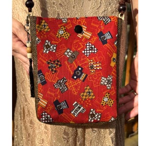 Puede incluir: Un bolso bandolera rectangular de tela roja vibrante con un patrón repetitivo de pequeños kimonos estilizados en varios colores y estampados geométricos. El bolso tiene paneles laterales texturizados en marrón oscuro y un cierre negro.