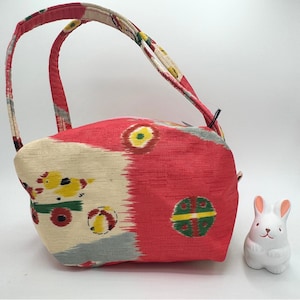 May include: A red and cream fabric handbag featuring a patterned design with yellow bird-like figures, green circular motifs, and two matching handles. Beside the bag sits a small, white, ceramic rabbit figurine with pink-tipped ears.