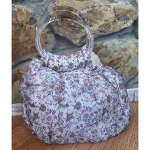 May include: A small, gathered fabric handbag featuring a light pink background with a dense floral pattern in shades of purple and dark brown. The bag is equipped with a single, rounded, transparent acrylic handle attached to the top.