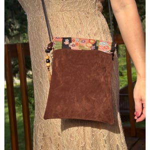 Puede incluir: Un bolso bandolera de ante marrón con cuerpo rectangular y solapa superior decorada con un estampado floral multicolor sobre fondo negro. El bolso incluye una correa negra fina y un adorno de borla lateral con cuentas de madera.
