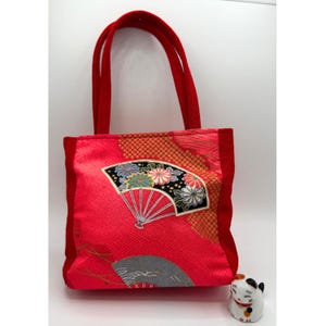 May include: Red tote bag with a Japanese fan design. The bag features a red background with a pattern and a black fan with floral accents. The handles and trim are a solid red. A small ceramic cat figurine sits beside the bag.