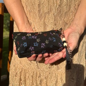 Puede incluir: Un estuche rectangular negro con cremallera que presenta un patrón repetitivo de pequeñas hojas de arce bordadas en varios colores. El estuche incluye una correa de muñeca de cadena metálica adornada con una cuenta blanca, una cuenta negra y una borla de tela negra.