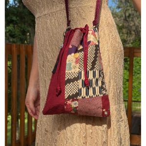 Può includere: Una borsa a sacchetto con coulisse caratterizzata da un design patchwork con vari motivi di ispirazione giapponese nei toni del bordeaux, crema, nero e oro. La borsa presenta un pannello laterale bordeaux testurizzato, una tracolla intrecciata e due lacci.
