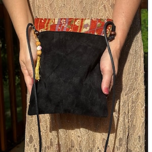 Puede incluir: Un bolso bandolera pequeño con cuerpo principal de ante negro y panel superior con estampado floral en rojo, dorado y morado. El bolso incluye una correa de cordón negro fino, cuentas de madera oscura y un dije de metal dorado con una borla amarilla.