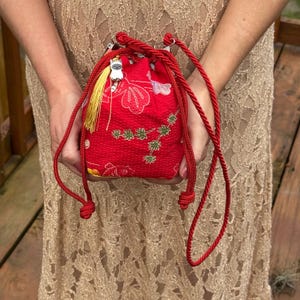 Op de afbeelding: Een rood trekkoordtasje van textuurstof met een wit en groen bloemenpatroon. De tas heeft lange rode koordhengsels en een kleine zilverkleurige kattenbedel bevestigd aan een gele kwast die aan de bovenste sluiting met trekkoord hangt.