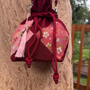 Puede incluir: Un bolso tipo saquito de color burdeos profundo con cierre de cordón, que presenta paneles triangulares de tela rosa con un estampado floral de flores de cerezo en blanco y dorado. Incluye una correa de cordón trenzado a juego y un colgante de borla.