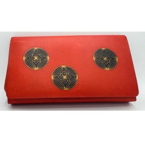 May include: A red clutch with three black and gold circular designs. The designs feature a floral pattern within a black circle, set against a vibrant red background. The clutch is rectangular and folded.