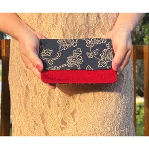 Puede incluir: Un bolso de mano rectangular con una sección superior azul marino oscuro con un patrón floral blanco y una sección inferior roja sólida con textura. El accesorio presenta un diseño horizontal limpio con un contraste claro entre paneles.