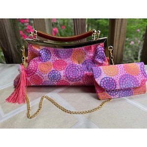 May include: A pink clutch purse and matching wallet set. The bags feature a repeating pattern of pink, purple, and yellow circles. The clutch has a gold frame with a wooden handle and a pink tassel. A gold chain strap is attached.