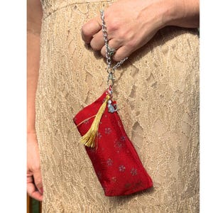 Puede incluir: Un pequeño bolso rectangular rojo con un sutil estampado floral, una borla dorada y un dije de gato plateado. El bolso está sujeto a una correa de muñeca de cadena de metal plateado con un cierre de mosquetón, creando un accesorio compacto.