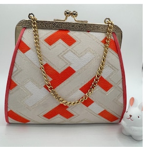 Peut inclure: Un sac à main structuré avec un motif géométrique orange vif, blanc et beige. Le sac possède une armature en métal doré avec un fermoir à pression et une bandoulière en chaîne dorée. Les bords sont garnis de tissu rose corail.