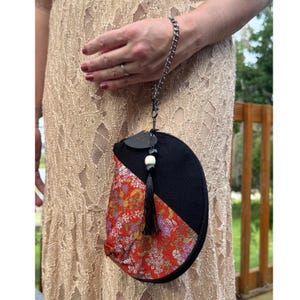 Puede incluir: Un bolso de mano semicircular con una sección superior de tela negra y una sección inferior de tela roja con un patrón floral multicolor. El bolso incluye una borla negra, una cuenta blanca y una correa de muñeca de cadena de metal plateado.