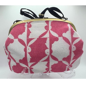 May include: A pink and white patterned clutch purse with a kiss-lock clasp. The fabric features a repeating design of stylized figures. The purse has a gold-toned frame, a black handle, and is displayed on a clear stand.