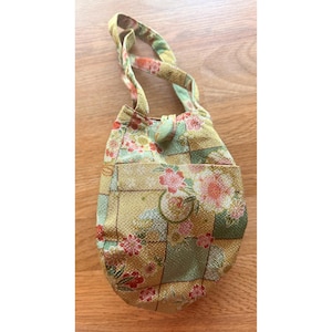 May include: A small, floral-patterned handbag with a rounded shape and a top handle. The bag features a patchwork design with squares of yellow, green, and pink floral fabric. A small pocket is visible on the front. The handle and button closure are green.