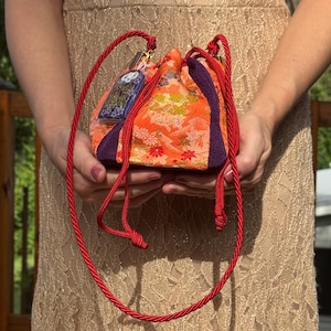 May include: A small drawstring pouch featuring a vibrant orange fabric with a floral pattern, accented by deep purple side panels. The bag includes long red braided cord handles and a small rectangular purple charm with white text attached.