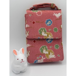May include: A pink fabric phone case with a flap closure and a small handle. The case features a repeating pattern of white rabbits, green and blue floral designs. A white ceramic rabbit figurine with pink ears is next to the case.
