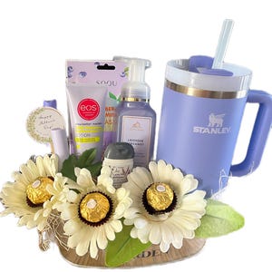 May include: A gift basket with a light purple Stanley mug, lotion, soap, and a small container of cream. The basket includes white flowers with gold-wrapped chocolates and a small round gift tag that says "Happy Mother's Day!"