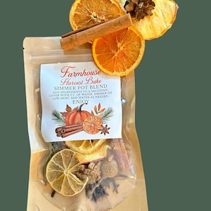 May include: A clear plastic pouch containing a Farmhouse Harvest Bake simmer pot blend. The label features text and a pumpkin illustration. Dried orange slices, a cinnamon stick, and a small pinecone are arranged on top of the pouch.