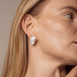 May include: A close-up of a white, irregular-shaped pearl stud earring. The earring is worn on a person's ear, with the focus on the lustrous, off-white pearl. The background is a soft, neutral tone, highlighting the earring's shape and colour.