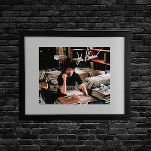 May include: A framed photograph featuring a person in a black shirt sitting amidst stacks of vinyl records. The image includes a white signature at the top and the text DO NOT COPY overlaid on the lower left side, all encased in a black frame.