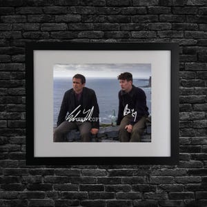 May include: Framed print featuring two men seated on a stone wall overlooking the ocean. The print is in a black frame with a white mat. The men are wearing dark clothing. The print includes signatures.