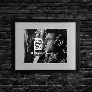 May include: Black and white framed print featuring a man in profile with a woman in the background. The print includes signatures and a playing card. The frame is black, and the background is a dark brick wall.