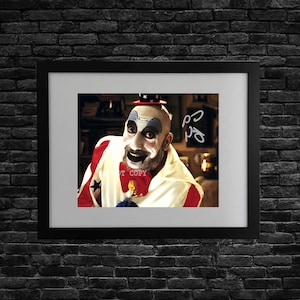 Puede incluir: Impresión enmarcada de un payaso con pintura facial blanca, un sombrero pequeño y un atuendo rojo y blanco salpicado de estrellas. El payaso tiene una pajarita roja pintada y una sonrisa siniestra. El fondo es un interior borroso.