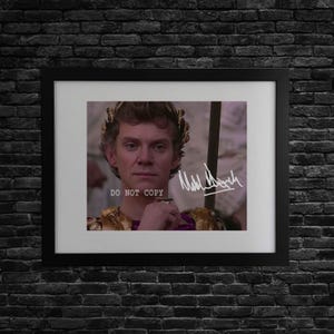 May include: Framed print featuring a portrait of a person wearing a gold laurel wreath and ornate clothing. The image includes the text "DO NOT COPY" and a signature. The frame is black, set against a dark brick wall.