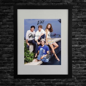 May include: A framed photograph featuring four individuals posing by a pool, with black ink signatures overlaid on the image. The group includes three people sitting on a ledge and one person sitting in front, all wearing casual clothing.