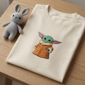 Könnte beinhalten: Cremefarbenes T-Shirt mit einem gestickten Bild eines grünen Alien-Charakters in einem braunen Mantel. Ein kleiner, gehäkelter Hase in Grau liegt neben dem gefalteten Shirt auf einer Holzoberfläche. Das T-Shirt ist ein Star Wars-Artikel.