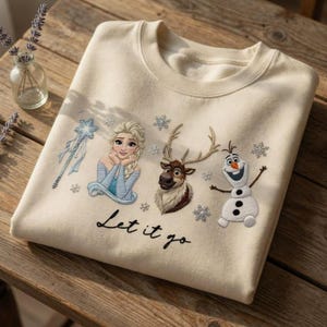 Può includere: Felpa color crema con personaggi ricamati del film Frozen: Elsa, Sven e Olaf, insieme a fiocchi di neve e una bacchetta. La frase "Let it go" è ricamata in nero.