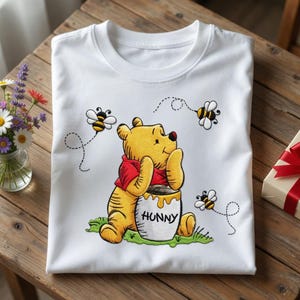 May include: White t-shirt featuring a cartoon illustration of Winnie the Pooh sitting next to a honey pot with the word "HUNNY" on it. The shirt also has bees flying around Pooh. A gift box with a red ribbon is on the right.