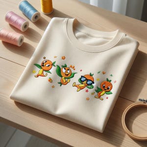 Puede incluir: Sudadera de cuello redondo color crema con un diseño bordado de cuatro personajes de dibujos animados naranjas con hojas verdes y gafas de sol. El diseño está rodeado de pequeñas flores y estrellas bordadas. Los carretes de hilo de bordar están en el fondo.