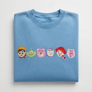 May include: A light blue sweatshirt featuring embroidered character faces from a popular animated film. The embroidered characters include a cowboy, an alien, a pink bear, a purple character, a cowgirl, and a pink pig.