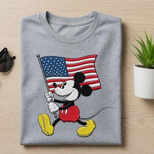 Könnte beinhalten: Ein gefaltetes graues T-Shirt mit einem Cartoon von Mickey Mouse, der eine amerikanische Flagge hält. Mickey trägt rote Shorts, gelbe Schuhe und weiße Handschuhe. Die Flagge hat rote und weiße Streifen und ein blaues Feld mit weißen Sternen.