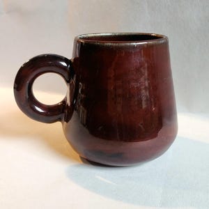 May include: A glossy, dark brown ceramic mug with a wide, rounded body and a prominent, circular handle. The glaze features subtle variations in tone, with a smooth, reflective surface and a slightly darker rim at the top opening.