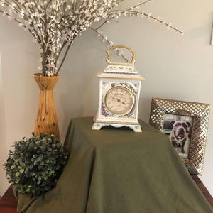 May include: A decorative white and gold clock with a round face, set on a green cloth. The clock has a gold handle and floral accents. A wooden vase with white branches and a framed photo are also visible.