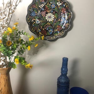 May include: A scalloped decorative plate with a black background and a colorful floral pattern featuring white, blue, green, and red painted details. The plate is displayed alongside a wooden vase with yellow flowers, a blue glass bottle, and a blue cup.